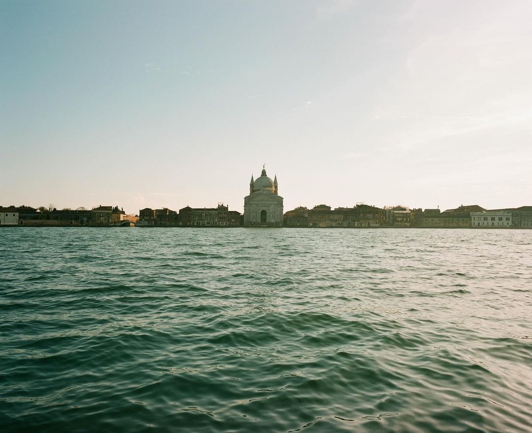 Venice cityscape featuring a tranquil teal waterway and classical architecture under a clear sky, captured in a wide landscape shot.