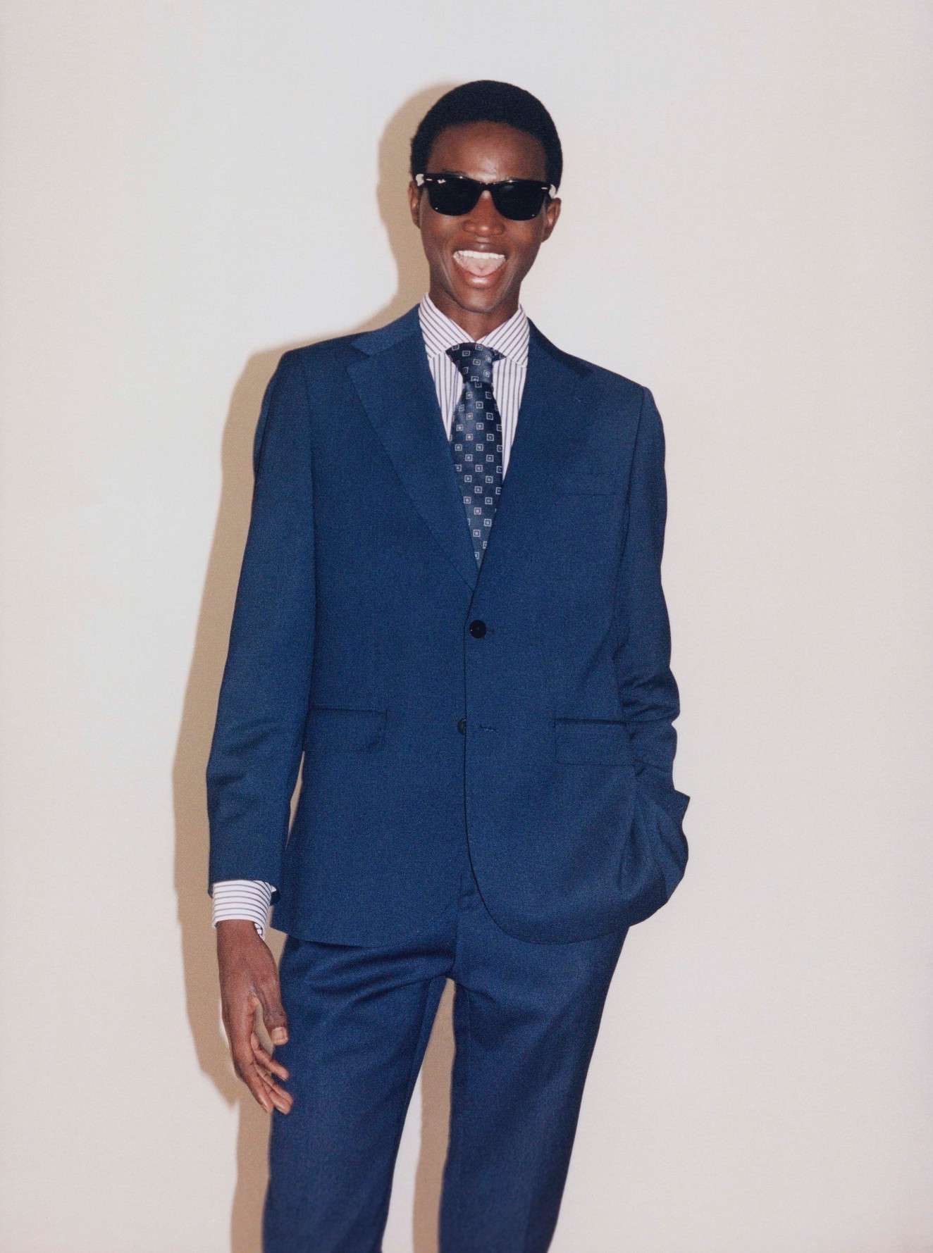 Full shot of a young adult male model with dark skin, wearing a blue suit and sunglasses, standing in front of an off-white background.
