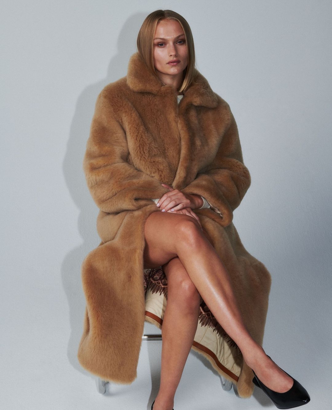 A fair-skinned adult woman sits wearing a long light-brown faux-fur coat and black heels in front of a plain white background.