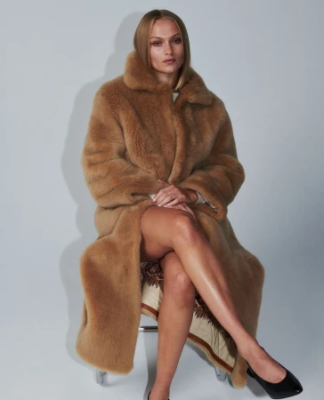 Model seated on a chair wearing a large brown faux fur coat against a neutral grey studio backdrop