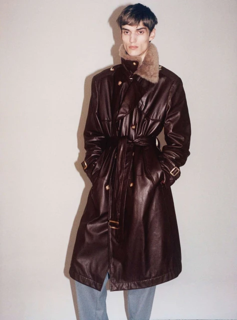 Man in a brown leather trench coat with a fur collar, posing indoors against a plain white wall in a fashion photography setting.