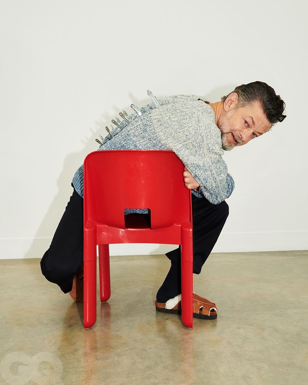 Mature man in gray sweater leans on red plastic chair with metal clips on back. He wears dark pants and brown sandals against white backdrop.