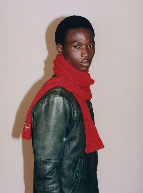 Studio portrait of a young adult Black man wearing a leather jacket and red knitted scarf, looking directly at the camera.