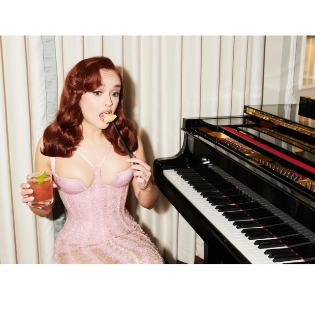 Red-haired woman in pink dress poses with cocktail and snack next to a piano inside, creating an elegant and stylish atmosphere.