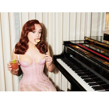 Red-haired woman in pink dress poses with cocktail and snack next to a piano inside, creating an elegant and stylish atmosphere.