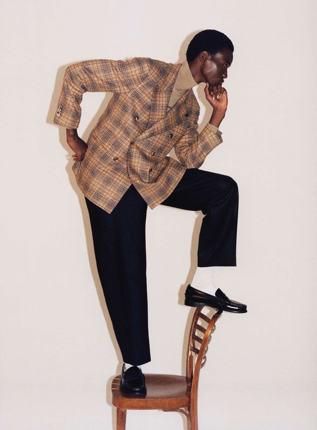 Fashionable Black man in a plaid blazer standing with one foot on a wooden chair in a photo studio setting.