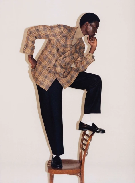 Fashionable Black man in a plaid blazer standing with one foot on a wooden chair in a photo studio setting.