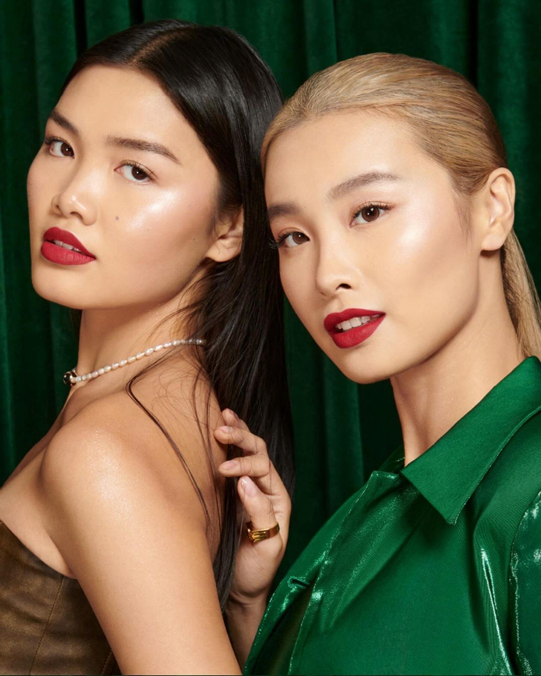 Two Asian women pose closely together in front of a green curtain, showcasing beauty and fashion with soft lighting and elegant makeup.