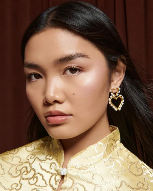 Close-up portrait of a woman with dark hair wearing a yellow and gold patterned top and gold heart-shaped earrings with pearls.