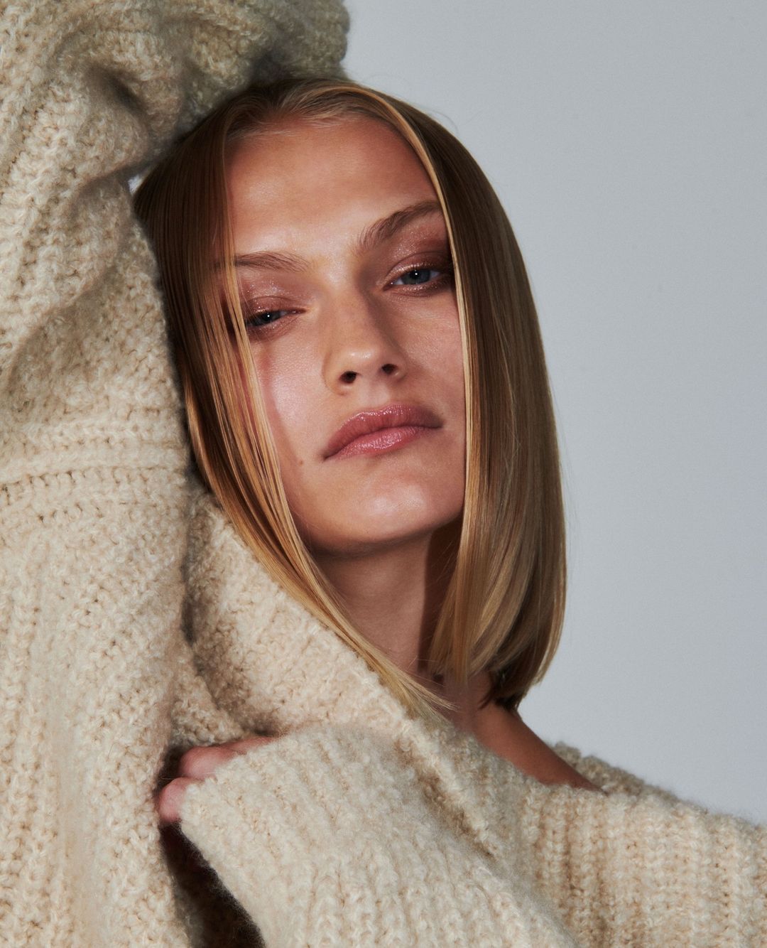 Close-up of a blonde adult female with fair skin and blue eyes wearing a beige sweater in a neutral studio environment.