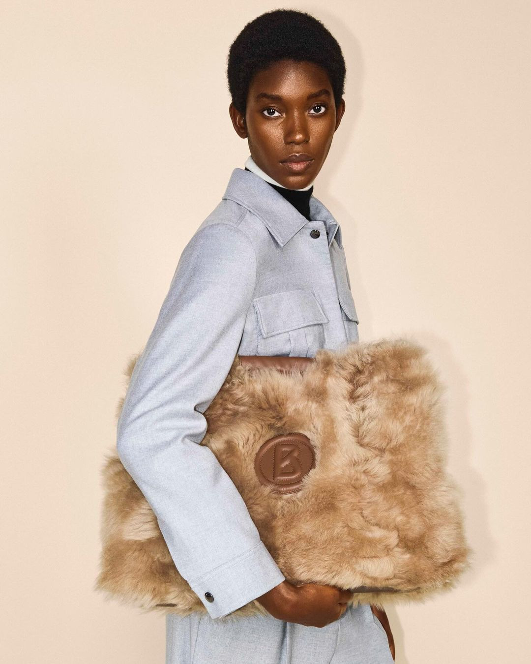 Woman in light blue jacket holding a large furry tote bag with a brown logo against a neutral backdrop in a studio setting.