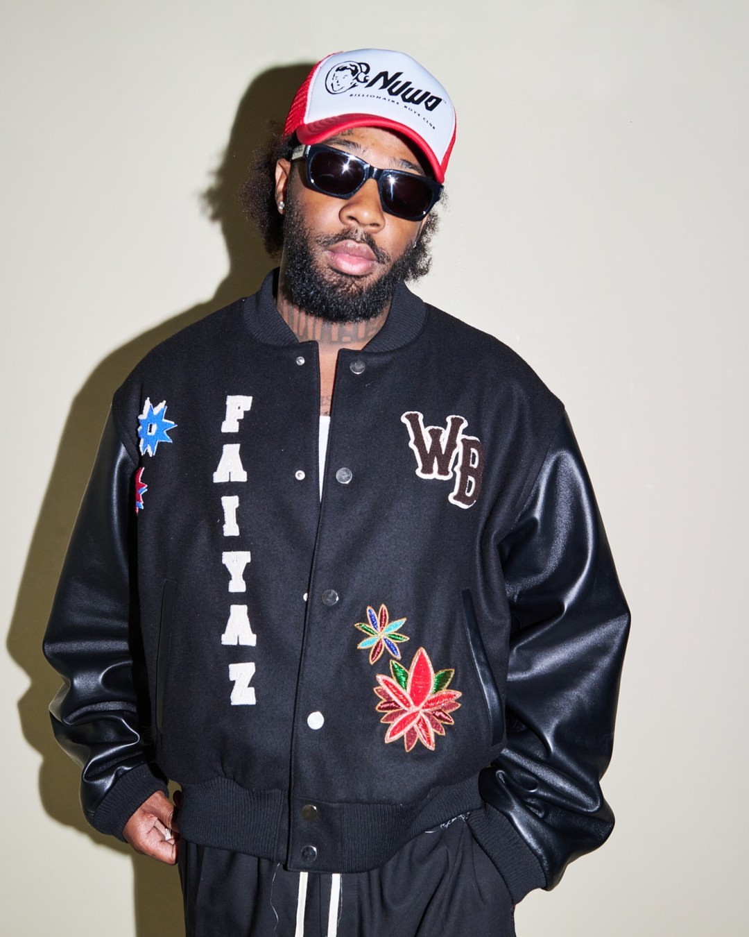 Man with a beard and sunglasses wears a hat and a black jacket with patches while posing indoors in front of a neutral backdrop.