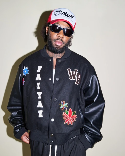Man with a beard and sunglasses wears a hat and a black jacket with patches while posing indoors in front of a neutral backdrop.