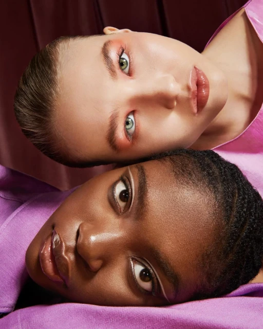 Close-up beauty shot featuring two women with contrasting skin tones, one light and one dark, lying side by side with makeup.