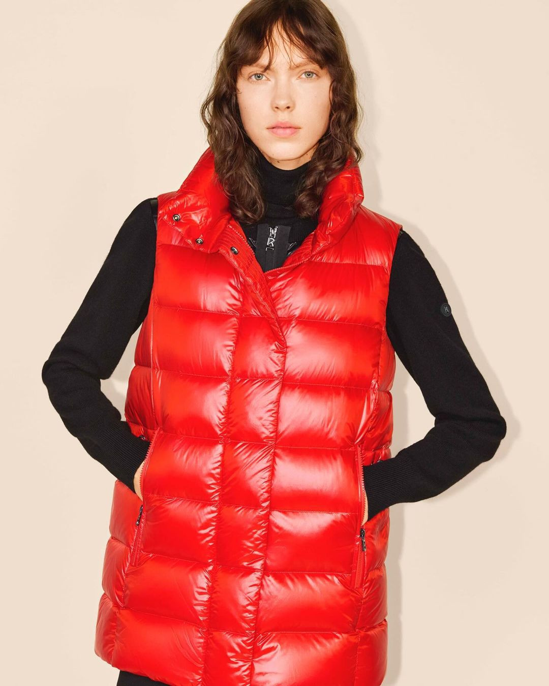 A young woman stands indoors wearing a vibrant red puffer vest and black turtleneck sweater. The medium shot is on a beige backdrop.