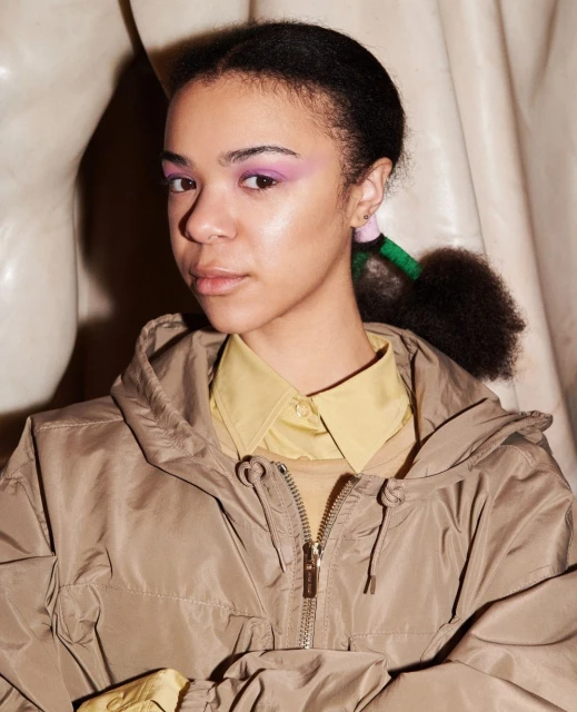 Close-up portrait of an adult woman with afro puffs wearing a tan hooded jacket and light yellow collared shirt, in a neutral indoor setting.