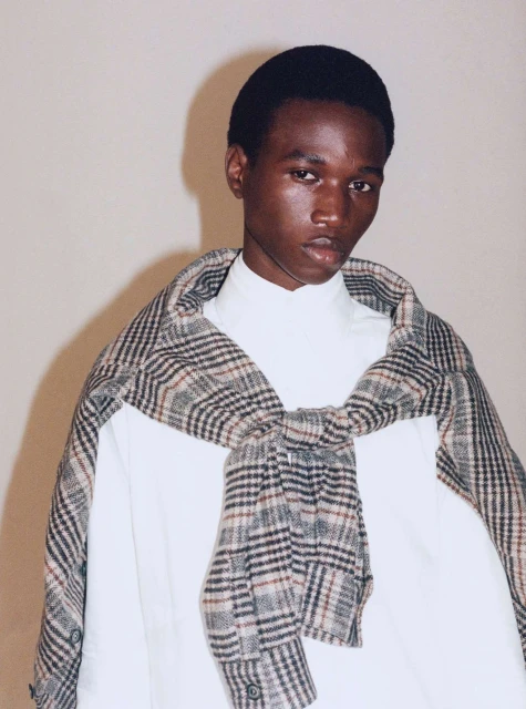 Headshot of a Black man wearing a white mock turtleneck and a plaid button-up tied around his shoulders against a neutral backdrop.