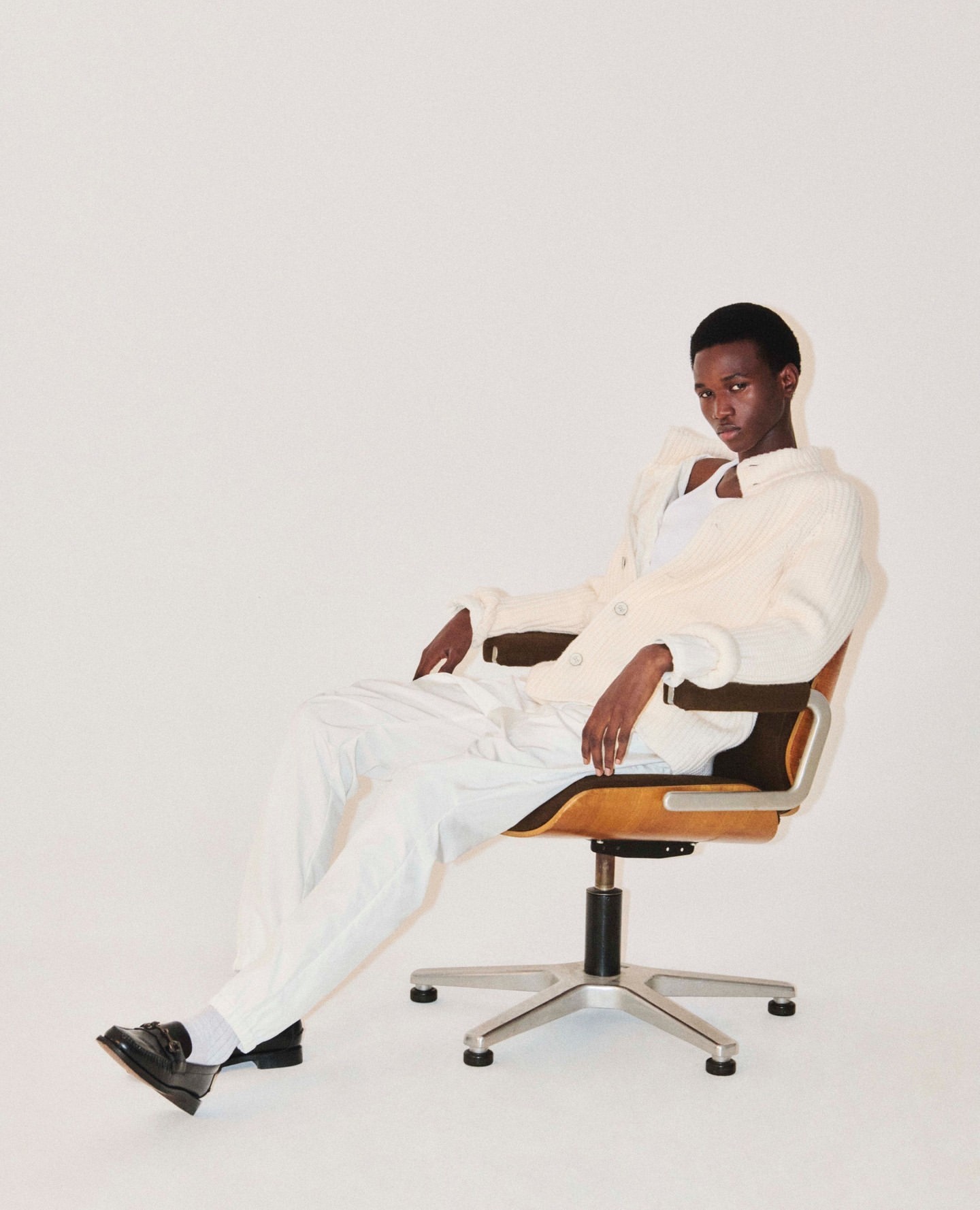 An adult male model poses in an off-white jacket and white trousers while seated in a rolling office chair in a bright, high-key studio.