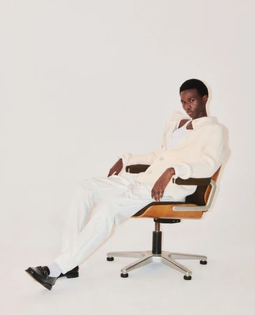 An adult male model poses in an off-white jacket and white trousers while seated in a rolling office chair in a bright, high-key studio.