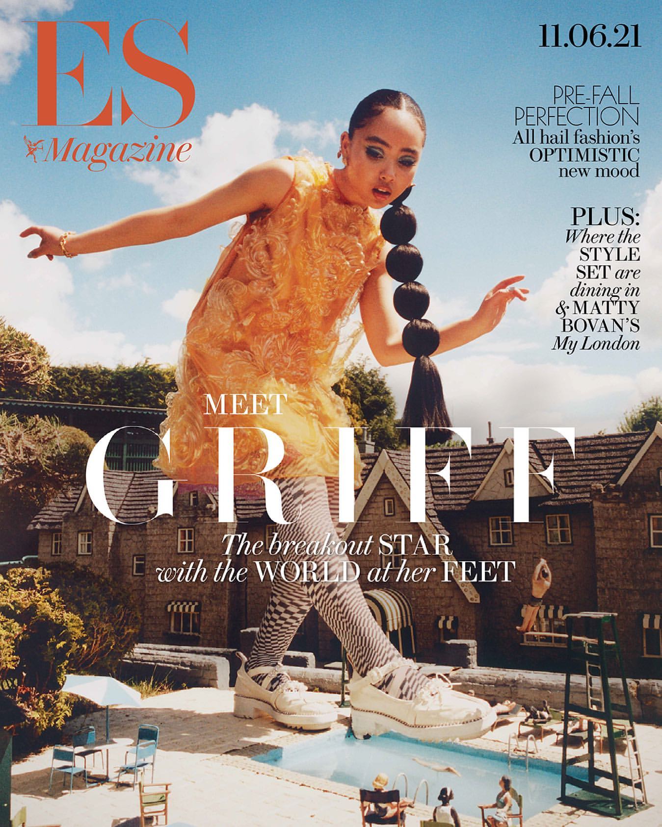 Magazine cover featuring woman in orange dress, sculptural hairstyle, oversized shoes, standing against miniature buildings and swimming pool.