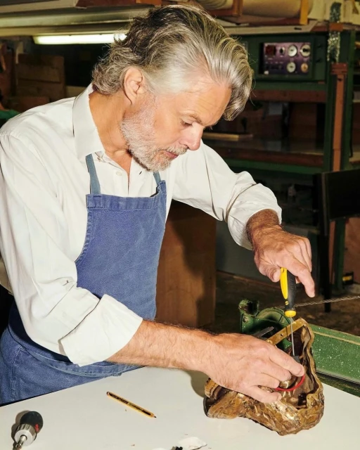 Mature man wearing apron works on a metallic object with a screwdriver in an indoor workshop, surrounded by tools.