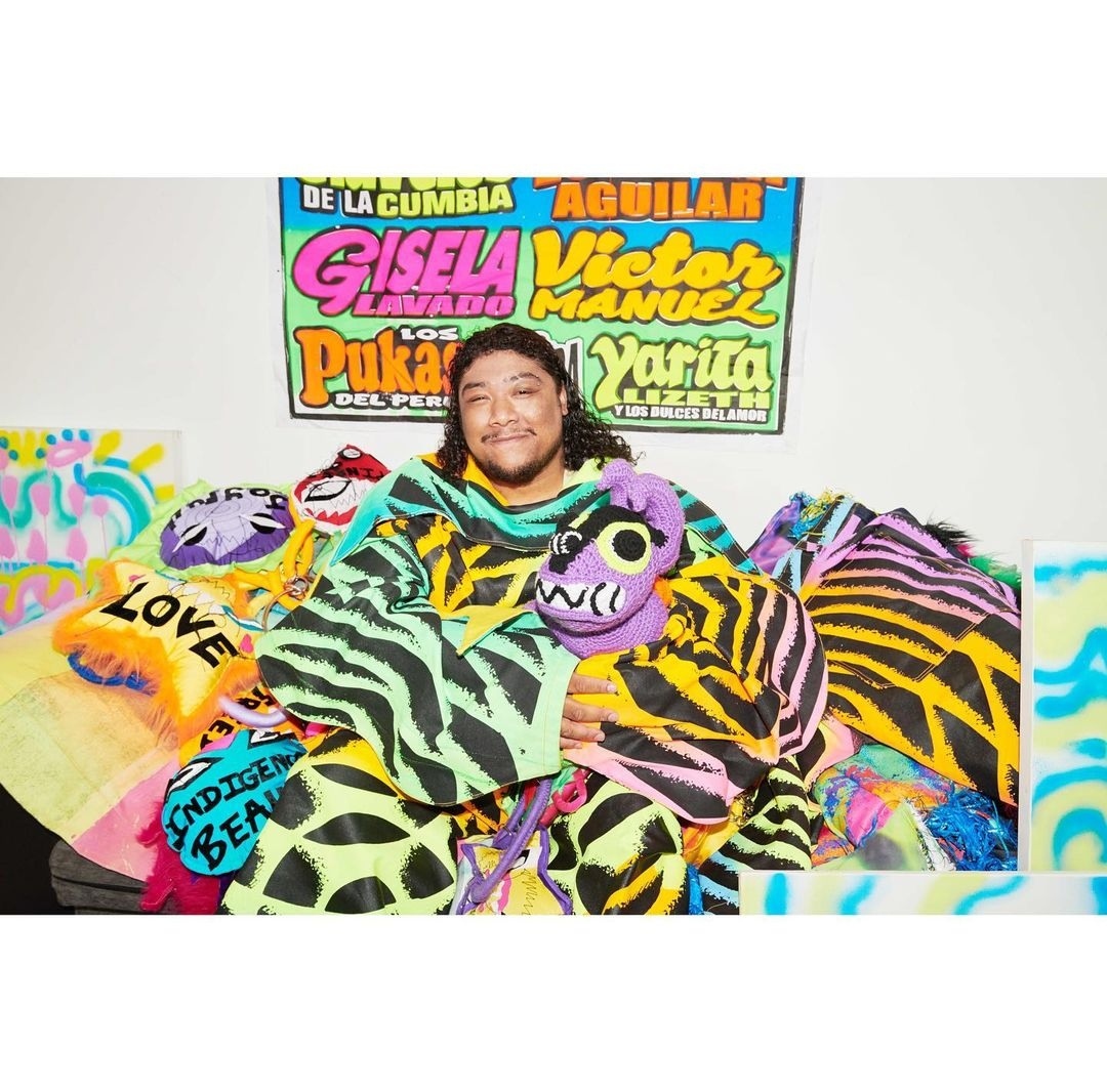 Portrait of a smiling person with long dark hair wearing a patterned poncho, hugging a plush animal. Colorful artwork in background.