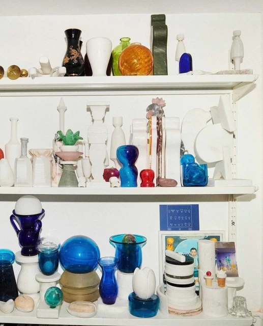 Collection of glass and ceramic vases and objects displayed on three white shelves against a white background in an interior setting.