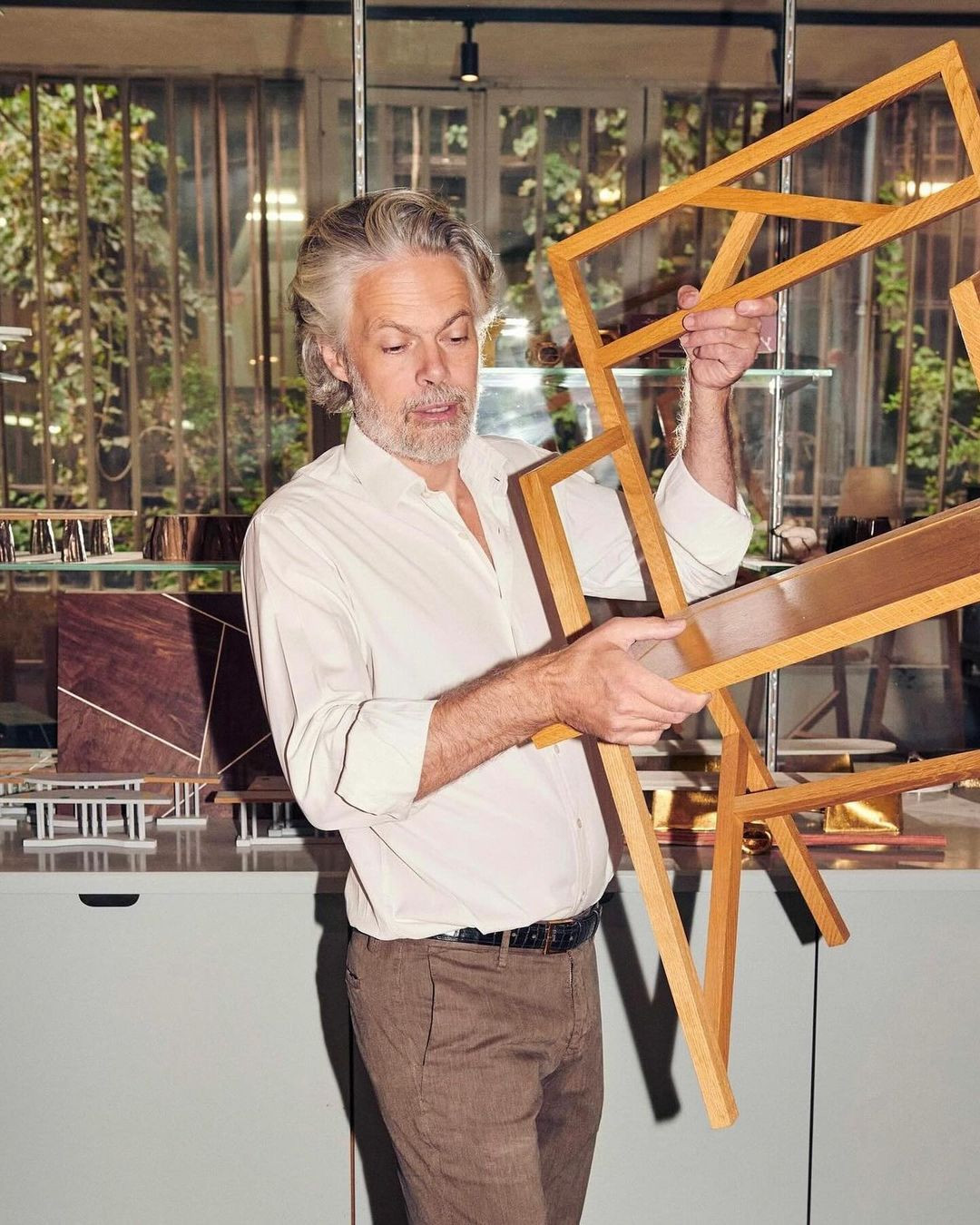 Mature adult man with grey hair holds wooden frames in a naturally lit indoor setting. He wears a white shirt and brown pants.