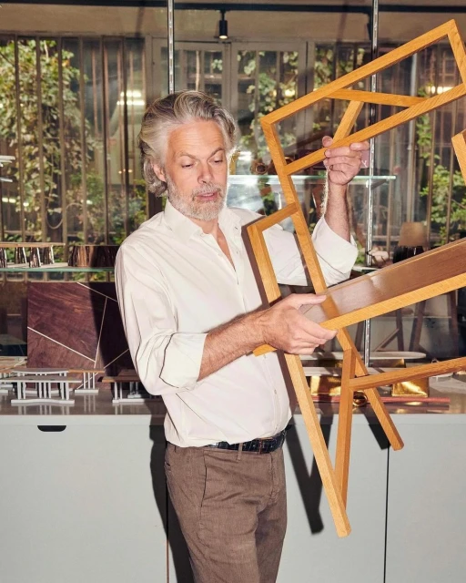Mature adult man with grey hair holds wooden frames in a naturally lit indoor setting. He wears a white shirt and brown pants.
