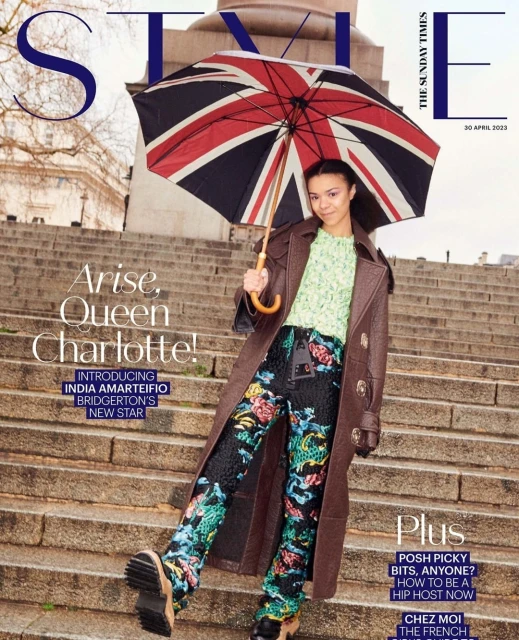Magazine cover featuring India Amarteifio, wearing a brown trench coat, patterned pants, and holding a Union Jack umbrella on stone steps.