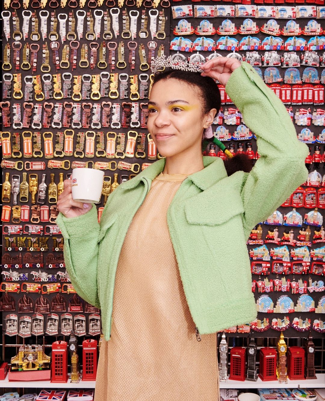 Young woman poses indoors with a white mug and a tiara, wearing a green jacket against a souvenir refrigerator magnet background.