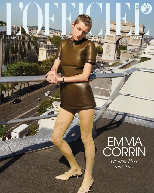 Emma Corrin poses on a rooftop in a brown leather dress and nude stockings against a Parisian cityscape on a magazine cover.