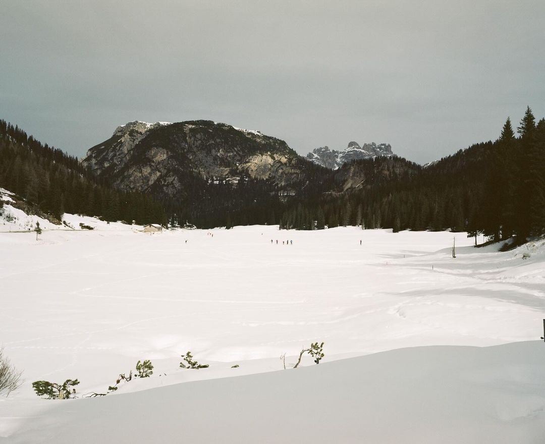 Mountain landscape with snow covered lake, evergreen trees and a group of people on the snow in the distance on a cloudy day.