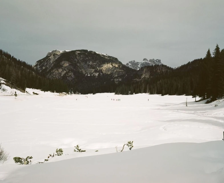 Mountain landscape with snow covered lake, evergreen trees and a group of people on the snow in the distance on a cloudy day.