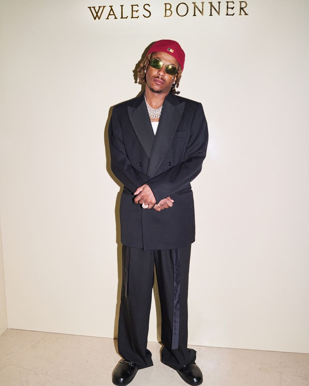 Man in a black tuxedo and red beanie standing in front of a wall with Wales Bonner branding in London.