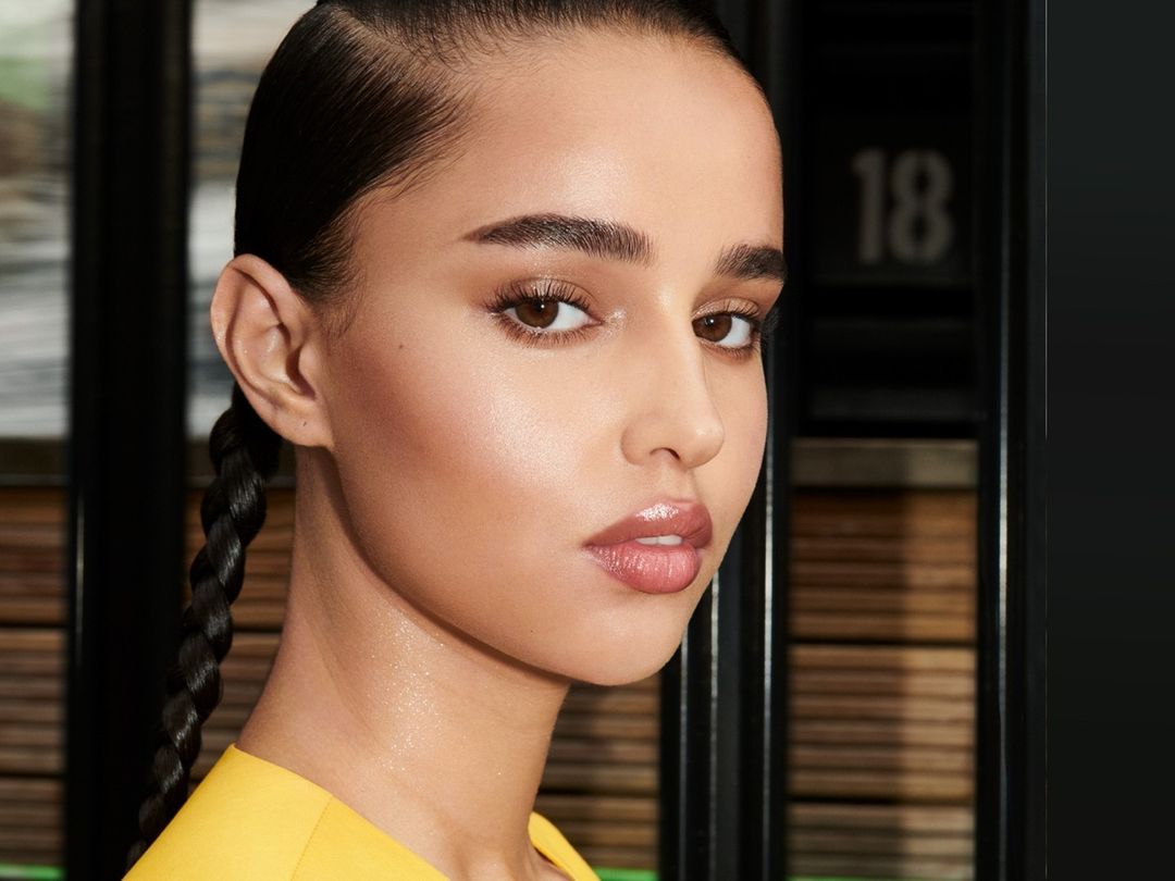 Close-up beauty shot of a young adult woman with brown eyes and black braided hair wearing a yellow top in a modern indoor setting.