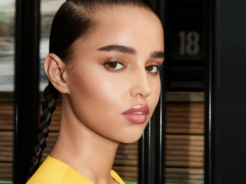 Close-up beauty shot of a young adult woman with brown eyes and black braided hair wearing a yellow top in a modern indoor setting.