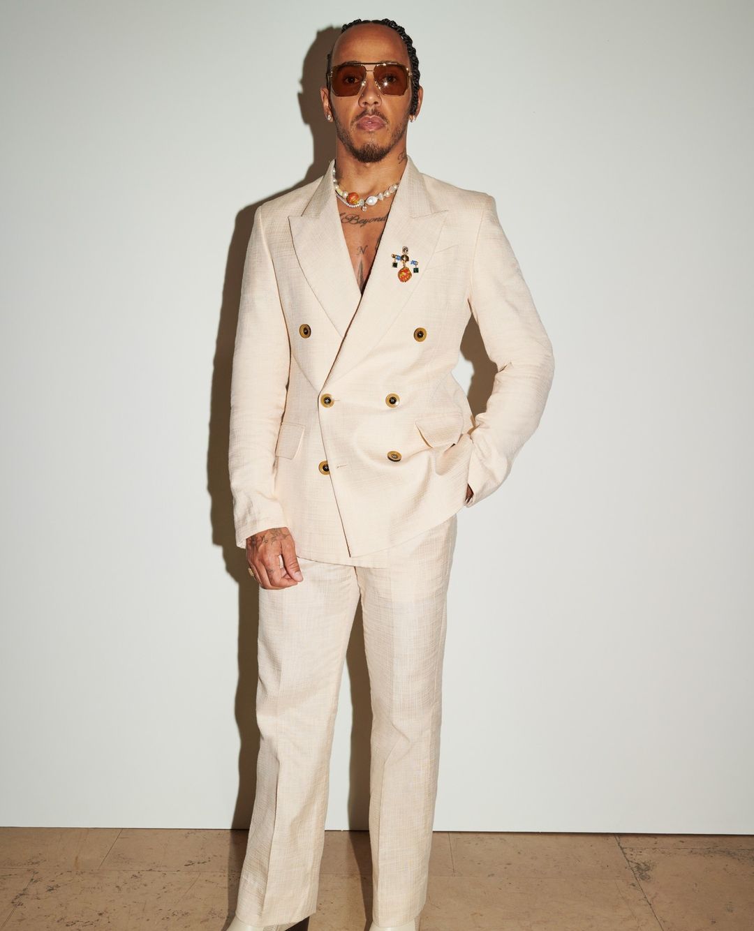 Portrait of Lewis Hamilton in a cream-colored suit with sunglasses and jewelry, posing against a white backdrop in a studio setting.