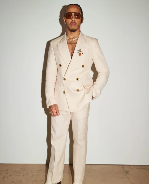 Portrait of Lewis Hamilton in a cream-colored suit with sunglasses and jewelry, posing against a white backdrop in a studio setting.