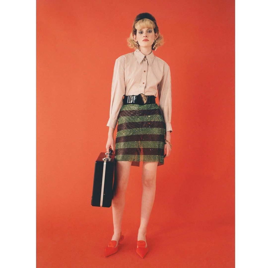 Adult female in a striped skirt stands against a red background holding a black briefcase while wearing red shoes and a button-down shirt.