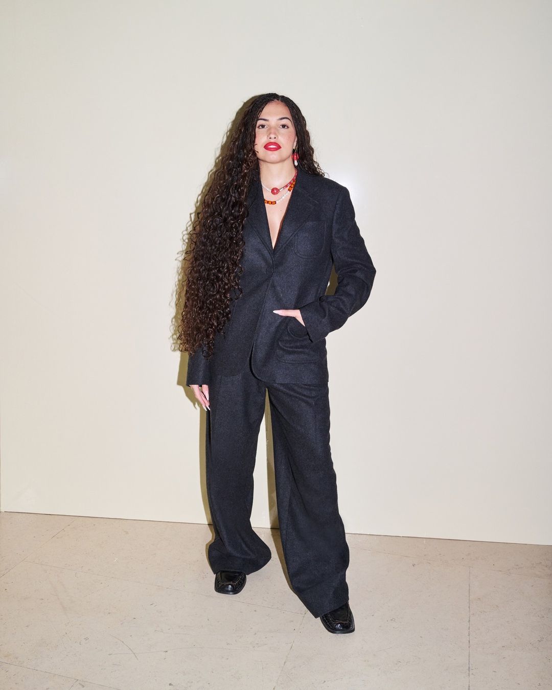 A curly-haired woman in a dark pantsuit poses indoors against a neutral wall, complemented by an amber necklace and earrings.