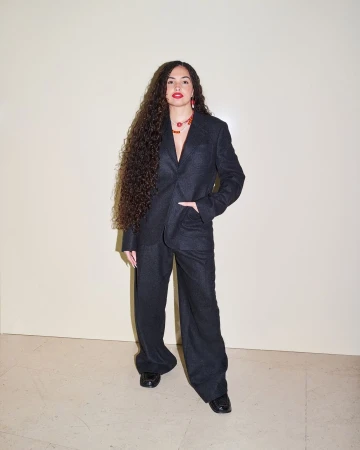 Woman with long curly hair wearing a dark grey suit and red lipstick against a plain wall