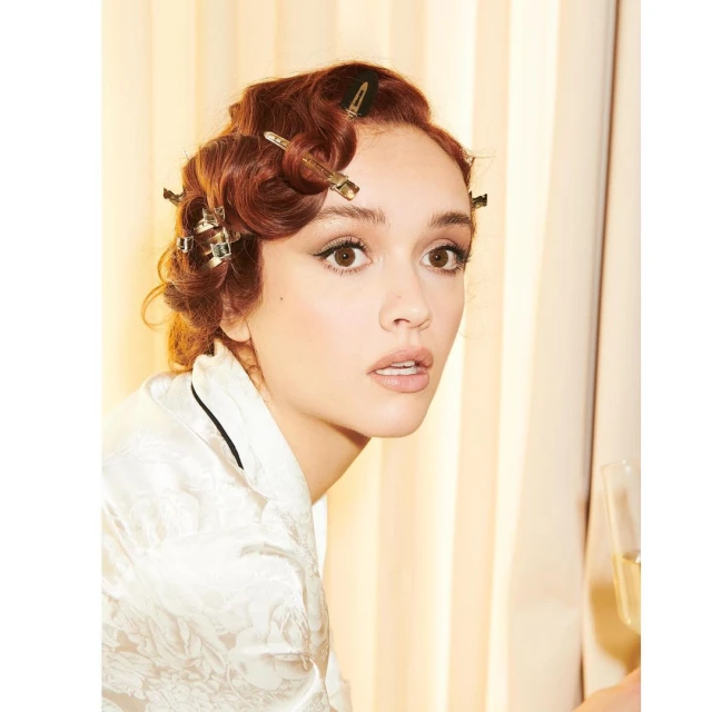 Close-up of a young woman with auburn hair styled in vintage waves with hair clips, wearing a silky patterned robe indoors.