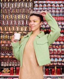 Woman in mint green textured jacket holding a white mug in front of a wall of souvenirs by Tung Walsh