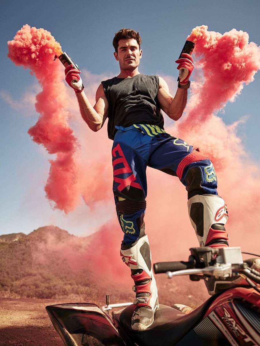 Adult male in action sports attire poses atop an all-terrain vehicle with red smoke flares against a clear sky and mountain backdrop.