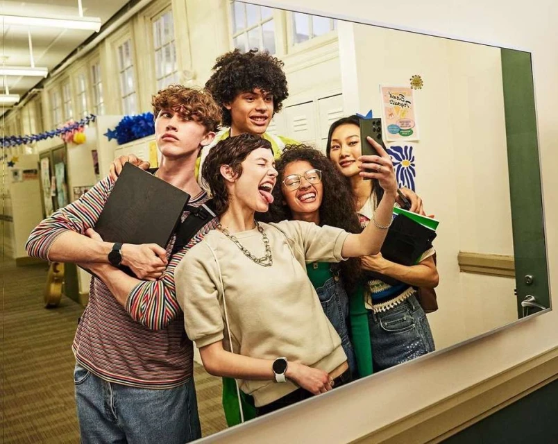 Group of five friends taking a selfie in a hallway, wearing casual clothing and smartwatches, capturing a candid moment with a smartphone.
