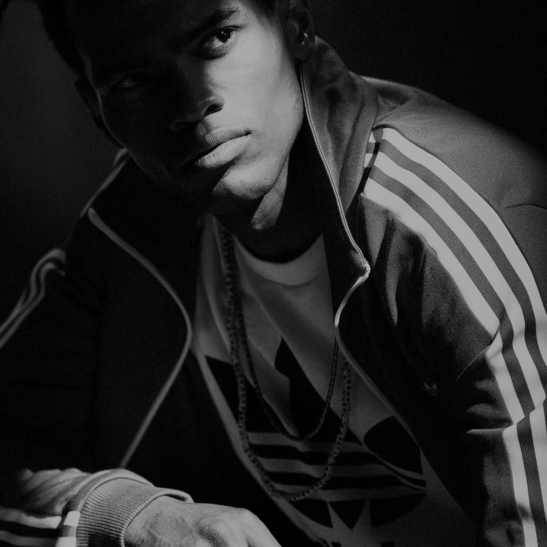 Monochrome portrait of a dark-skinned man wearing a tracksuit jacket and chain necklace, glancing at the camera in a dimly lit indoor setting.