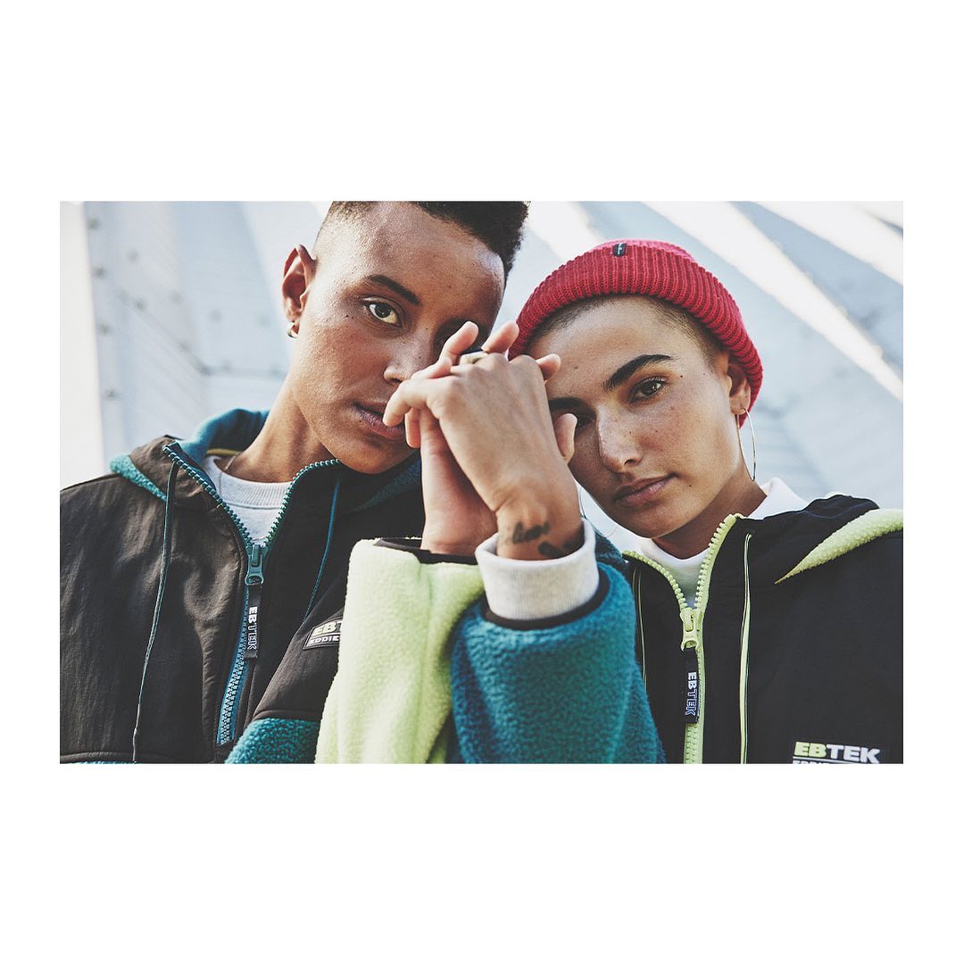 Two people pose close together outdoors, wearing athletic jackets and beanies, against a gray textured wall with soft natural light.