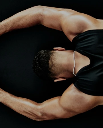 Top-down portrait of a muscular man in a black t-shirt, with his arms stretched out against a dark backdrop, emphasizing his physique.