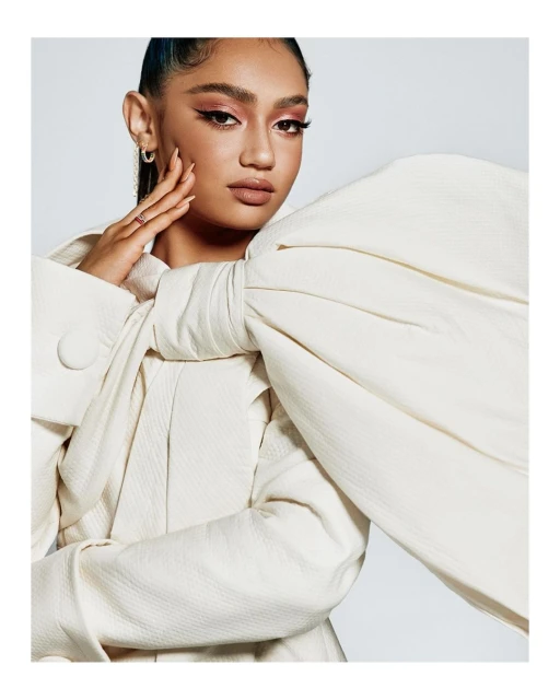 A woman poses in a studio wearing a cream-colored dress with a large, dramatic bow, showcasing beauty makeup and stylish fashion.